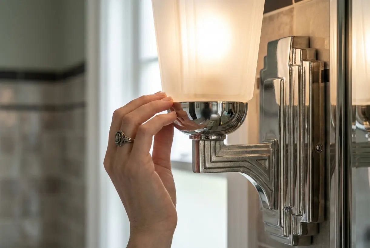 Art Deco bathroom wall sconce with geometric chrome detailing being adjusted in elegant bathroom setting.
